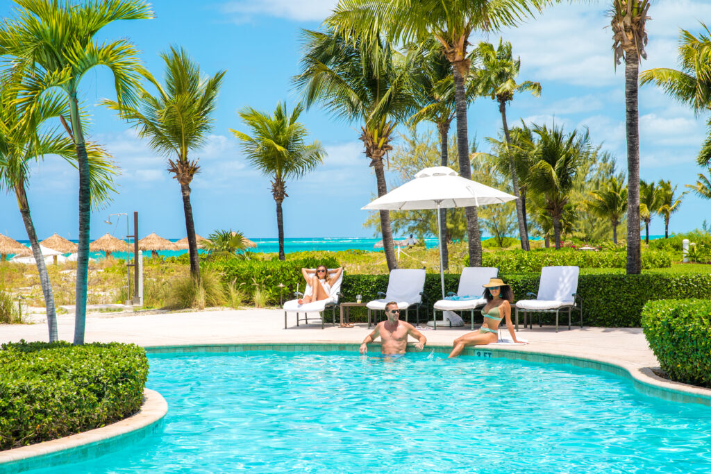A woman in a lounge chair by the pool with a man and woman relaxing in the pool.